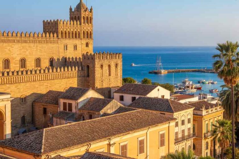 Medieval stone fortress overlooking a harbor with boats, tan buildings, and a palm tree against a blue sea horizon at sunset/calm light.