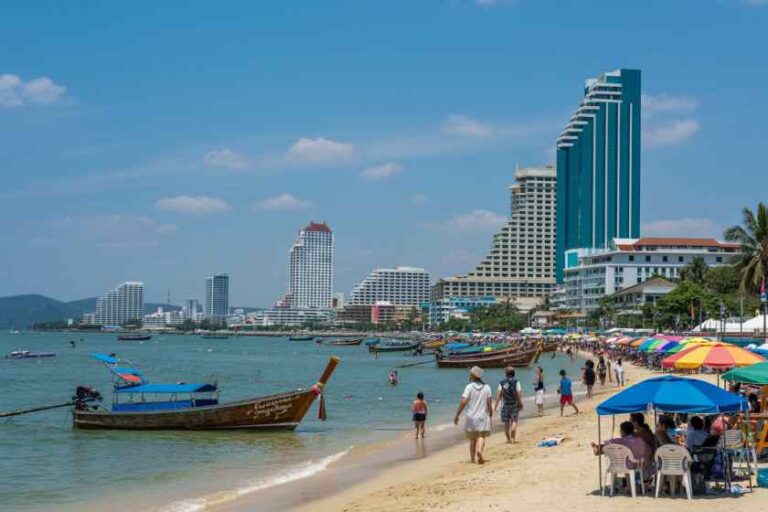 Beach scene with wooden long-tail boats near shore, colorful umbrellas, and tall hotel buildings along a sunny coastline.