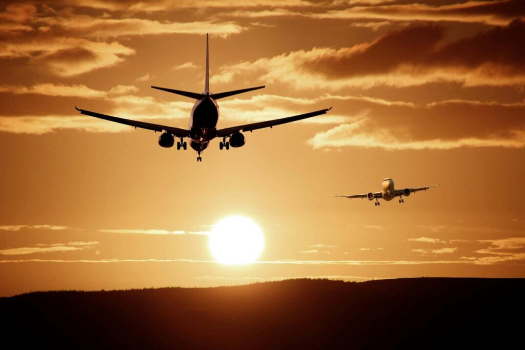 Two commercial airplanes approach for landing against an orange sunset, with the larger plane in the foreground and a smaller one in the distance.