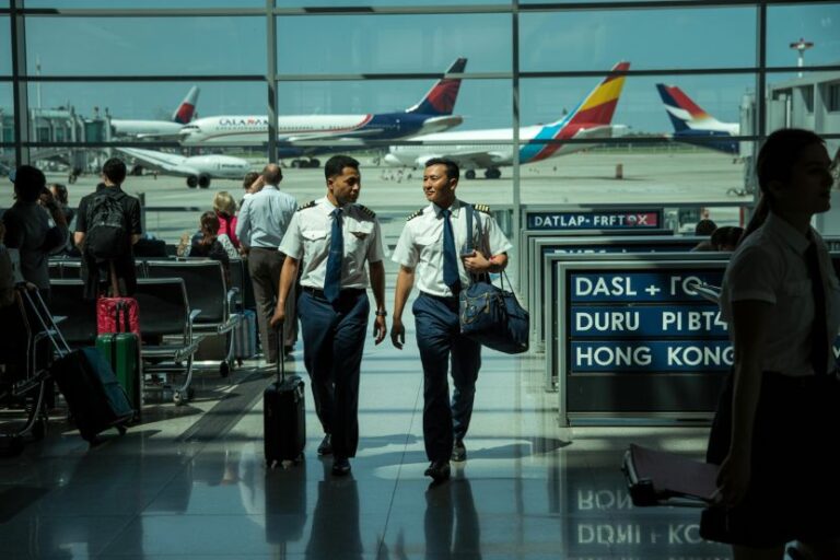 Two pilots in uniform walk through a bright airport terminal with luggage as planes taxi outside through large windows.