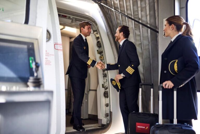 Two pilots in uniform shake hands at the doorway of an airplane as a crew member with luggage watches nearby.] ,