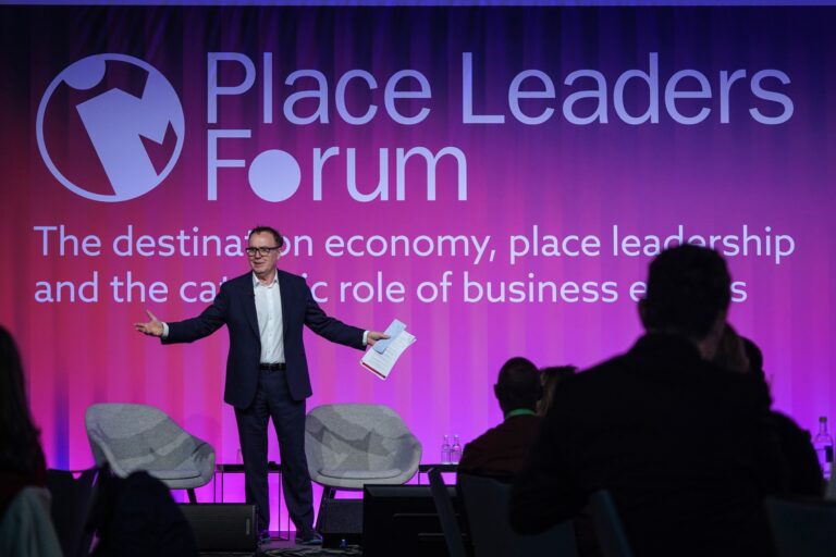 Speaker in a dark suit on stage at Place Leaders Forum, gesturing with papers in hand against a purple backdrop with large text
