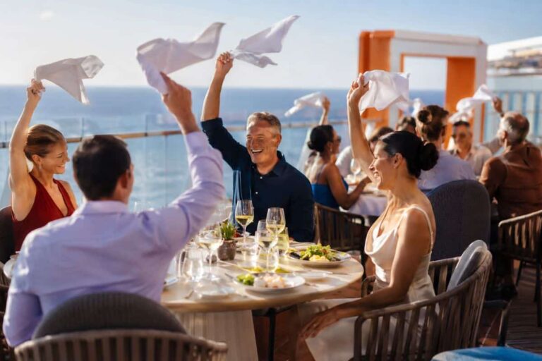 Group of adults at a seaside outdoor dinner on a cruise deck, raising white napkins in celebration with wine glasses on the table and the ocean in the background.