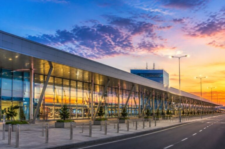 Modern glass-fronted airport terminal with angled support columns along a roadway at sunset, blue-pink sky reflecting on windows.