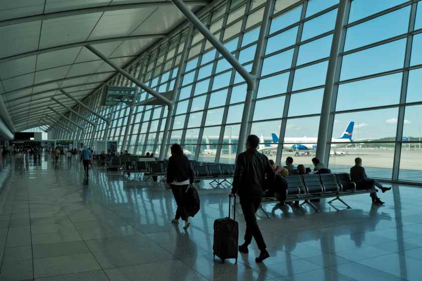 Airport terminal with large glass walls; travelers walk by with rolling suitcases, planes visible outside the gates.