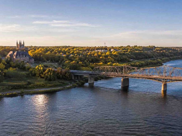 A river with a metal truss bridge spanning it, church-like buildings and green trees on the riverbank bathed in warm evening light.