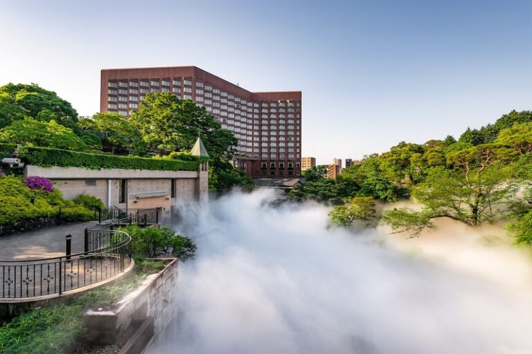 Brick high-rise building overlooking a misty river valley with lush green trees and a curved stone walkway in the foreground, daytime sky clear.