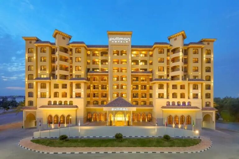 Front view of a multi-story Pullman hotel at dusk, warmly lit with arched ground-floor windows and a central entrance canopy.