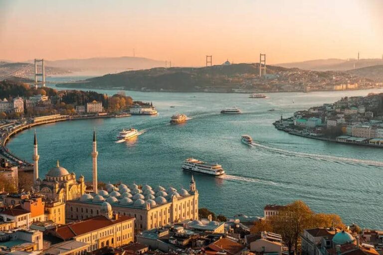 Aerial view of Istanbul at sunset with the Bosphorus Strait, ferries sailing, and a domed mosque with minarets in the foreground.