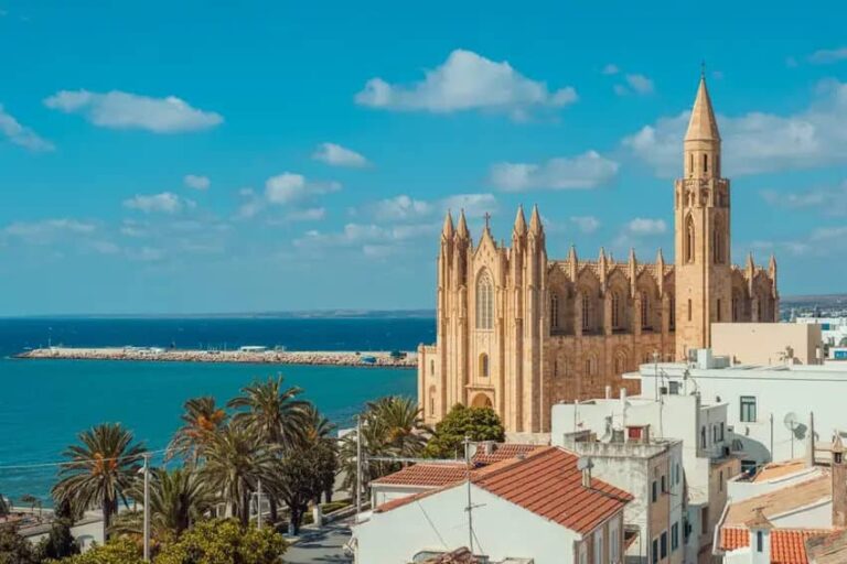 Coastal town with a large Gothic-style stone church overlooking the blue sea and palm-lined promenade.