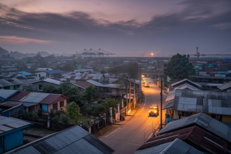 Dusk over a small town: a narrow street with parked cars and modest houses, with a container port and cranes on the horizon.