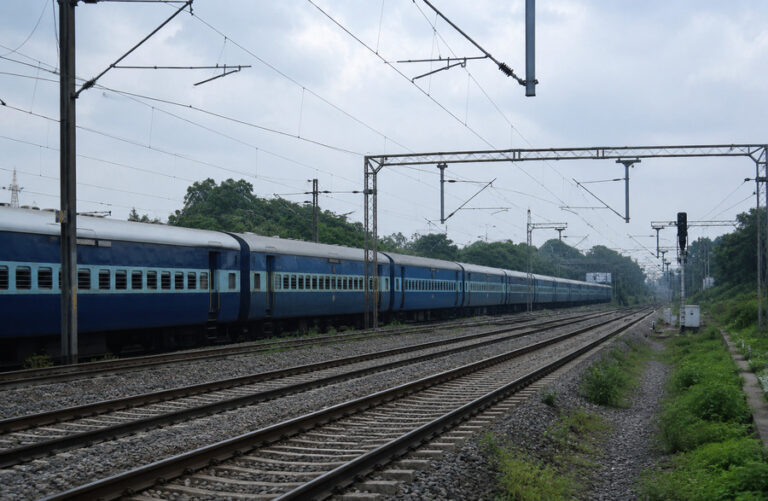 Long blue passenger train stretches along parallel tracks under an overhead electric line system, with green vegetation on both sides.