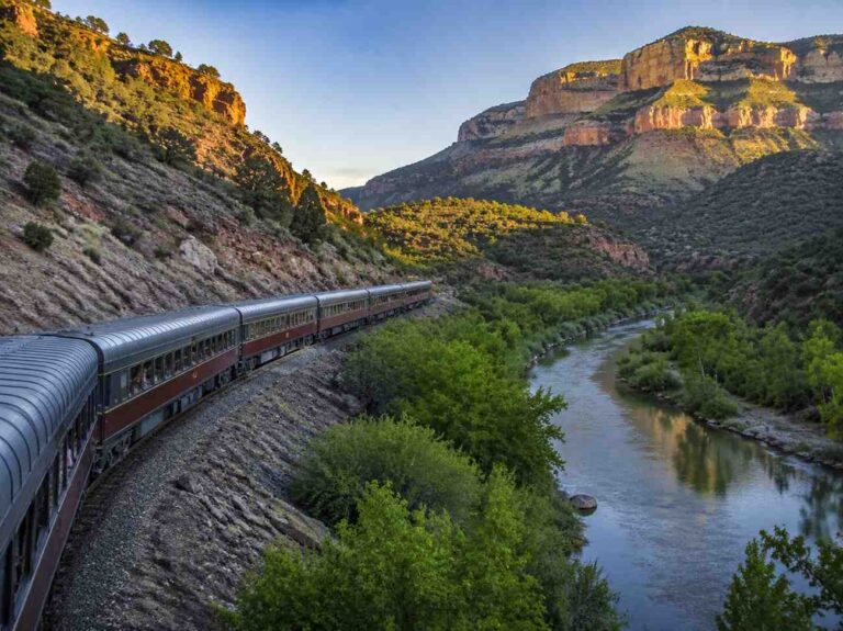 Passenger train winds along a curved track through a canyon, with a river on the right and red rock cliffs nearby.