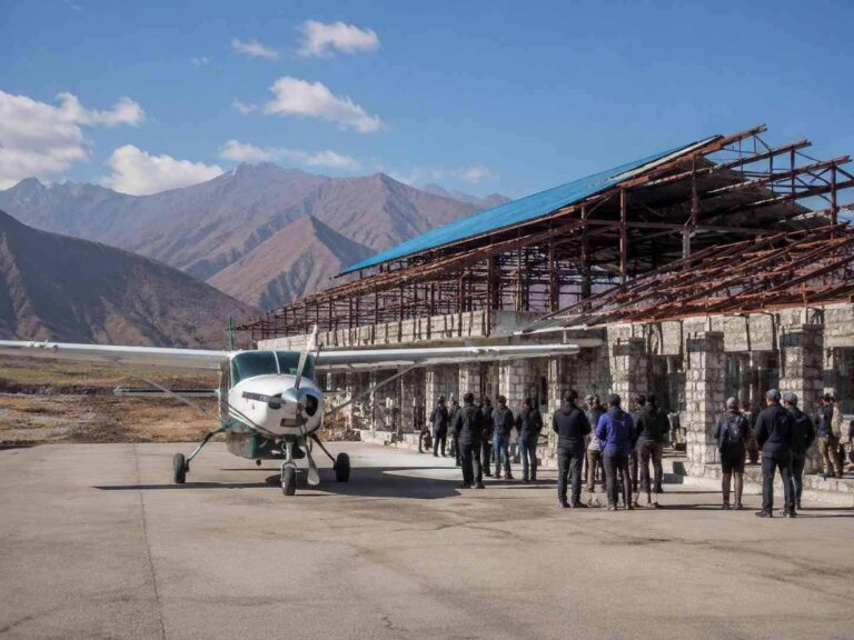Small propeller plane on a tarmac next to a stone hangar under construction, with a group of people in jackets nearby and rugged mountains in the background.