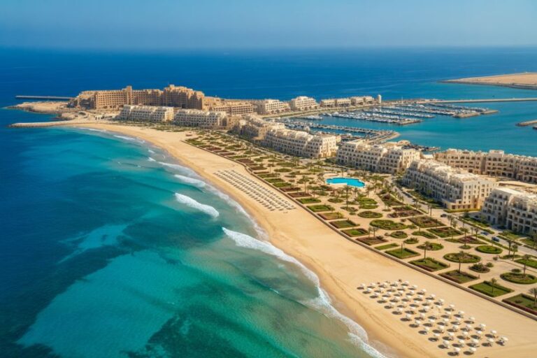Aerial view of a luxury beachfront resort with white buildings, a sandy beach, rows of sun loungers, palm trees, and a marina along turquoise water.