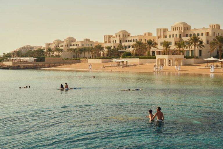 Resort shoreline with white villa-style buildings, palm trees, and people swimming in calm turquoise water by a sandy beach at sunset/early morning light.