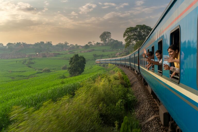 Passengers lean out of a bright blue train as it travels along a curved track through green fields at sunset.