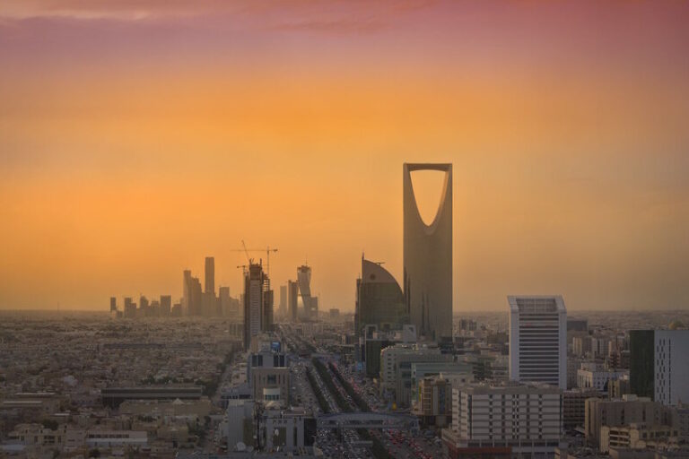 Riyadh skyline at sunset, featuring the Kingdom Centre with its distinctive arch, and a row of midrise buildings along a highway.