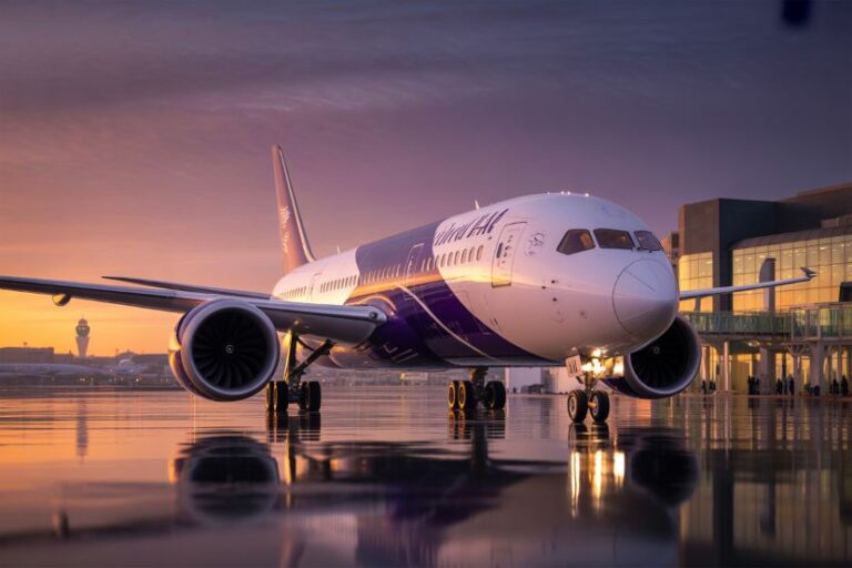 Virgin Atlantic wide-body airliner on a wet runway at sunset, with terminal building in the background and reflections on the tarmac.
