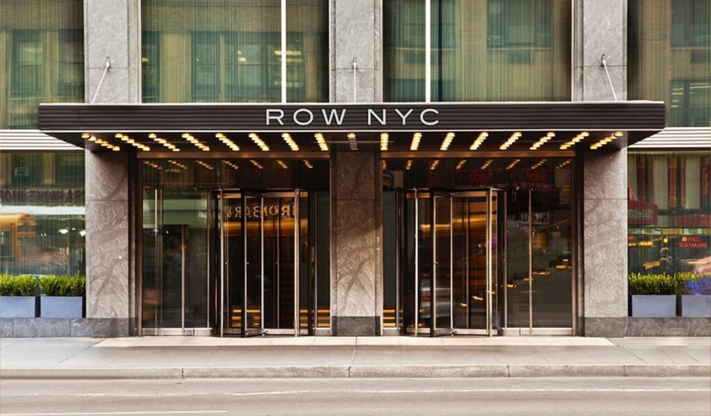 Hotel entrance with glass doors beneath a black marquee reading ROW NYC, illuminated by string lights above the doors in a modern building façade.