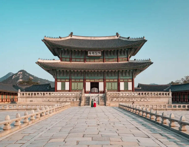 Two people in colorful hanbok stand before a large traditional Korean palace gate with a stone courtyard under a clear blue sky.