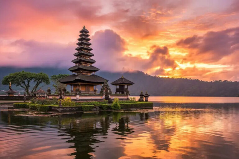 Balinese temple complex on a small island in a lake at sunset, with a tiered pagoda and calm, mirror-like water reflecting the sky.