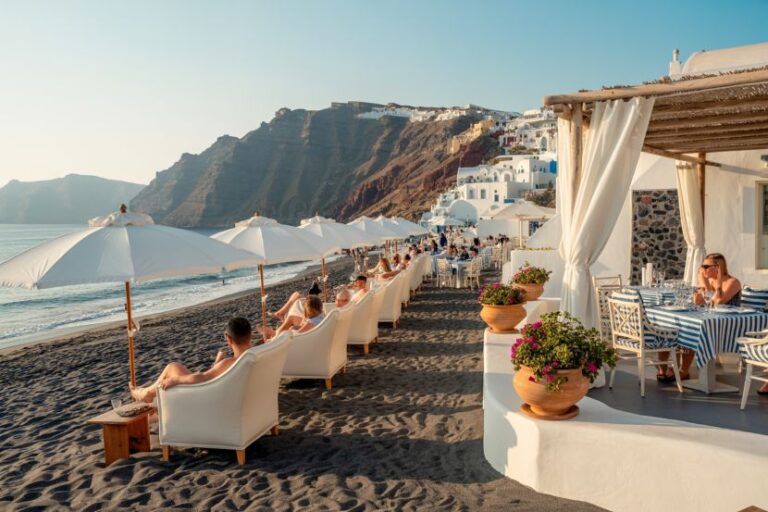 Beachfront restaurant with white lounge chairs under umbrellas along a dark sand beach; cliffs and white buildings rise in the background.
