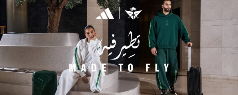 Ad banner for a sportswear brand showing a man in a green tracksuit and a woman in white, with rolling suitcases and the text 'MADE TO FLY' over a modern lobby backdrop.