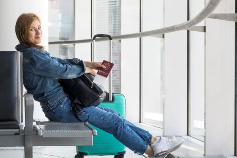 Woman sits in an airport terminal with a turquoise rolling suitcase, holding a passport and a small bag, smiling at the camera.