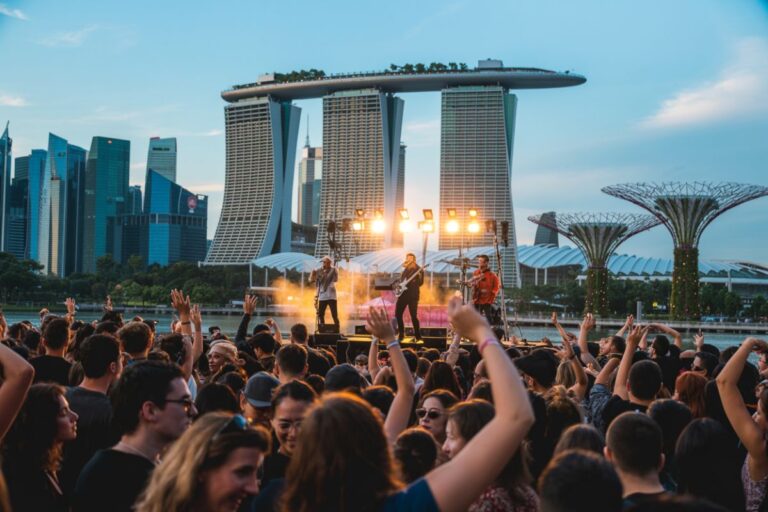 Band performs on an outdoor stage by the water as a large cheering crowd watches, with Singapore's skyline in the background.