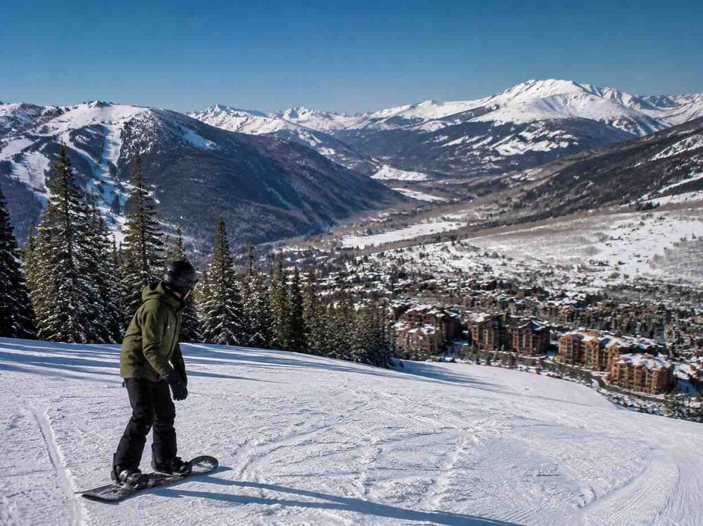 Snowboarder wearing a green jacket gliding down a snowy slope with a town and snow-covered mountains in the background.