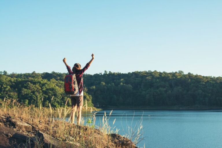 Person with a red backpack standing on a rocky lakeshore, arms raised in celebration with forested hills in the background.