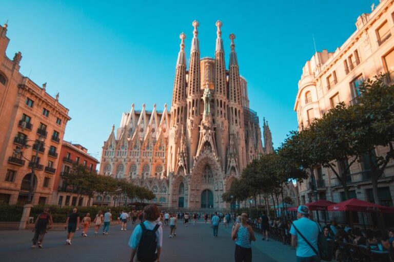 Sagrada Família cathedral in Barcelona, with tourists walking in the sunny plaza and trees framing the scene