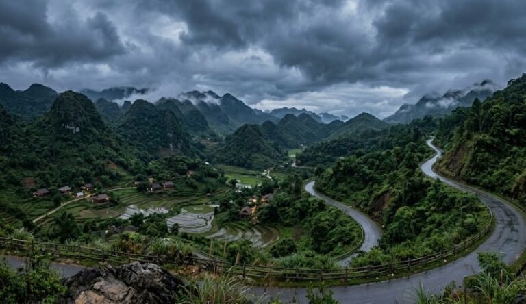 Winding road cuts through a green valley with terraced rice fields and small houses, surrounded by misty, cloud-covered mountains.
