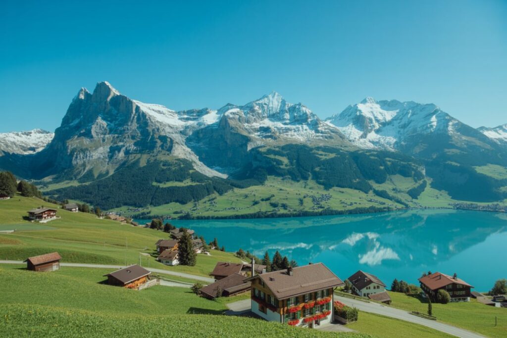Alpine village with wooden houses on green hills beside a turquoise lake and snow-capped mountains under a blue sky.