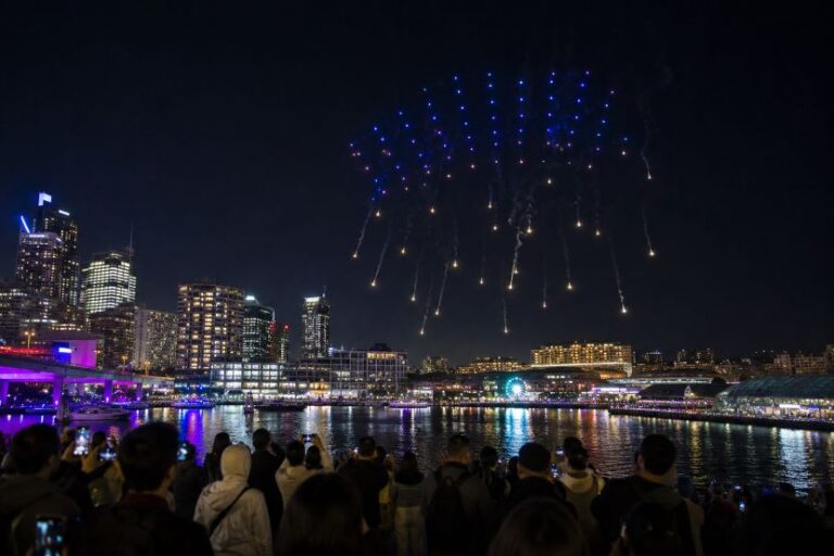 Night scene of a harbor city at night with blue and yellow fireworks exploding over the water, spectators in the foreground watching and filming.