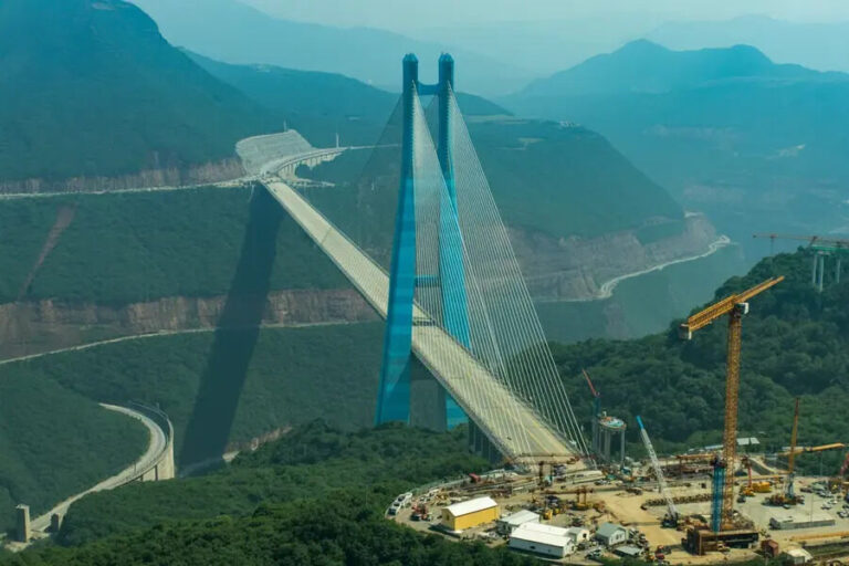 Blue cable-stayed bridge under construction spanning a deep valley, with cranes and a busy construction site at the base.