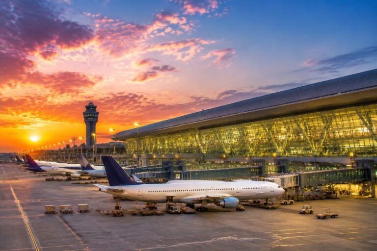 Sunset over an airport tarmac with planes at gates, a glass-walled terminal, and a control tower in the distance.