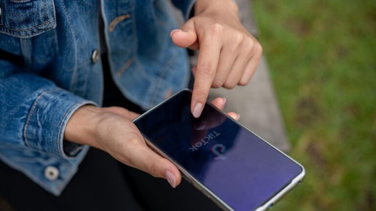 Close-up of hands holding a smartphone outdoors, with the TikTok app open on the screen and one finger tapping it.