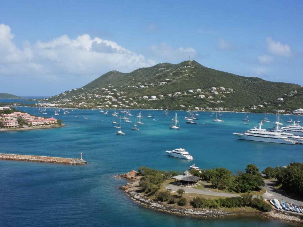 Harbor with numerous sailboats and yachts on turquoise water, green hills in the background and a blue sky above.
