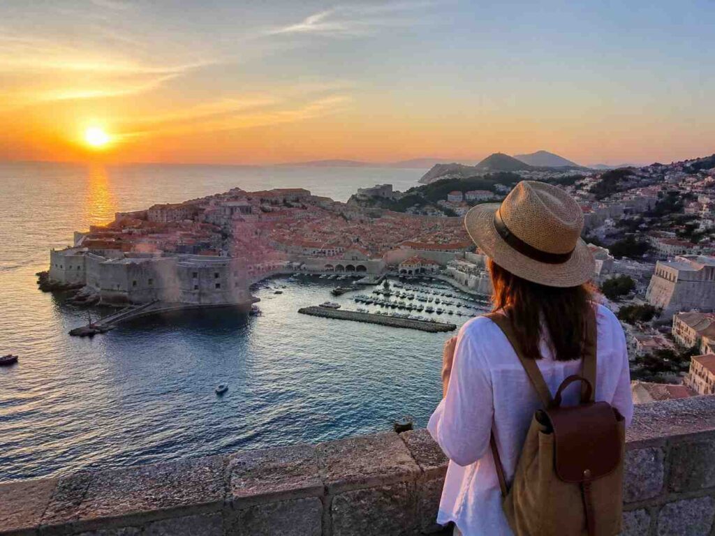 Traveler with a straw hat and brown backpack stands on a stone wall, watching a sunset over a historic coastal fortress and harbor town.