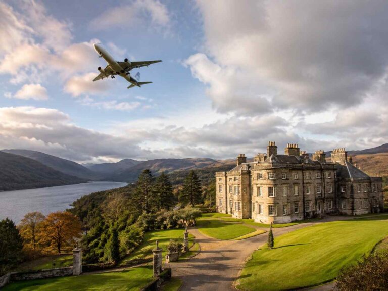A large stone castle on a manicured lawn beside a river valley, with an airplane flying low overhead in a partly cloudy sky.