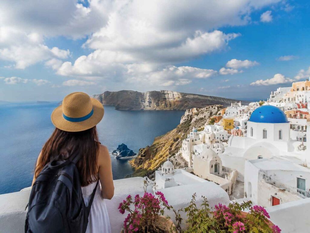 Woman in a straw hat and backpack gazes at Santorini's white buildings, blue-domed church, and the sea.