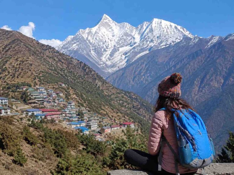 Person with a blue backpack sits on a ledge, looking at a snow-capped mountain village below.