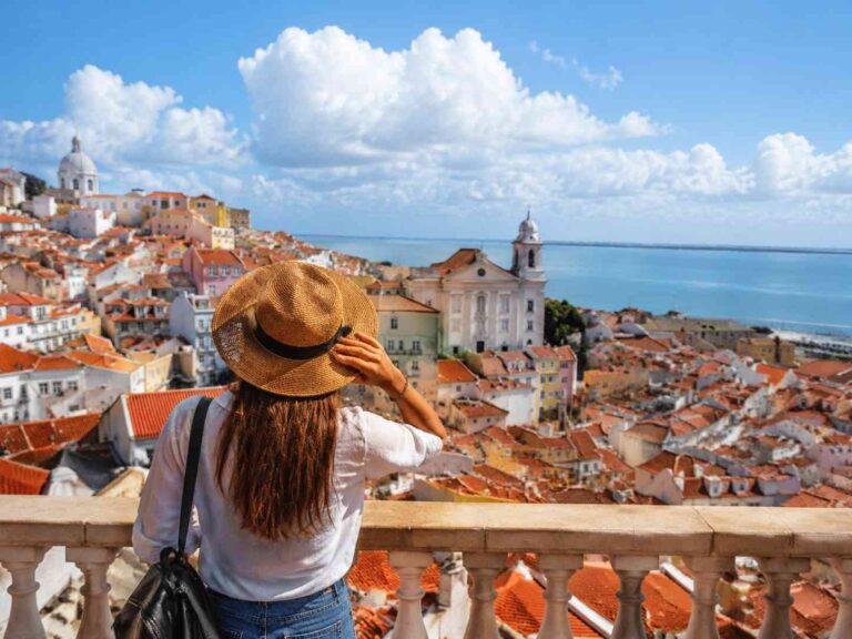 Woman wearing a straw hat stands at a railing, overlooking a hillside town with orange-tiled roofs by the sea under a blue sky