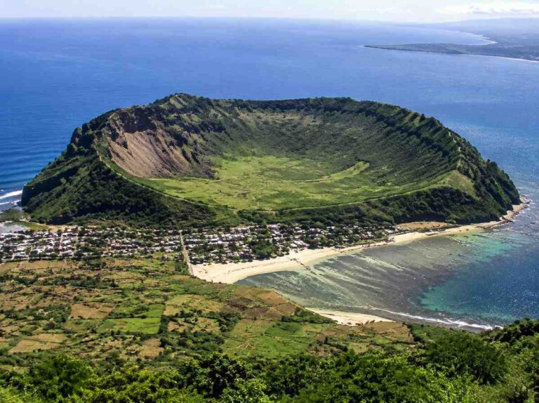 Circular volcanic crater with green inner crater on a coastal island, rim surrounded by blue ocean and a small beach town at the shore.