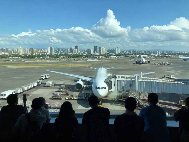 Silhouetted travelers watch a white Airbus at a gate, with jet bridge attached and airport equipment on the tarmac.