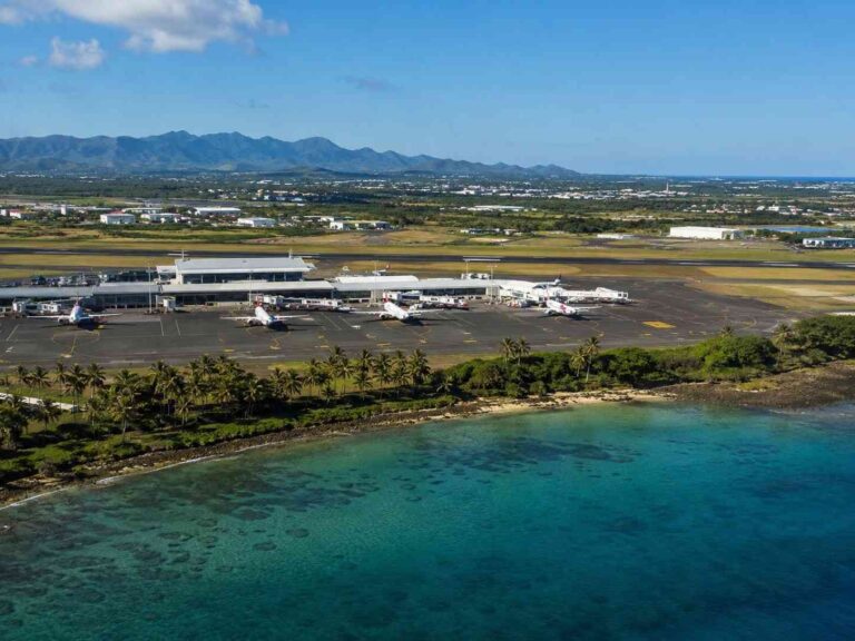 Coastal airport with a terminal and small passenger planes parked at gates, palm trees along the shoreline and turquoise water in the foreground.