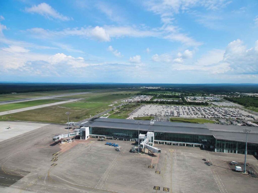 Aerial view of a large airport terminal with jet bridges, surrounding parking lots, and runways under a bright blue sky.