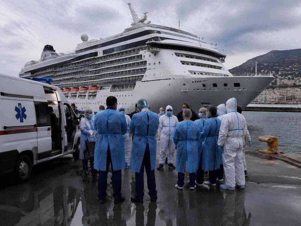 Medical team in blue gowns and white protective suits stand in a circle near a large cruise ship at a harbor, with an ambulance visible nearby.
